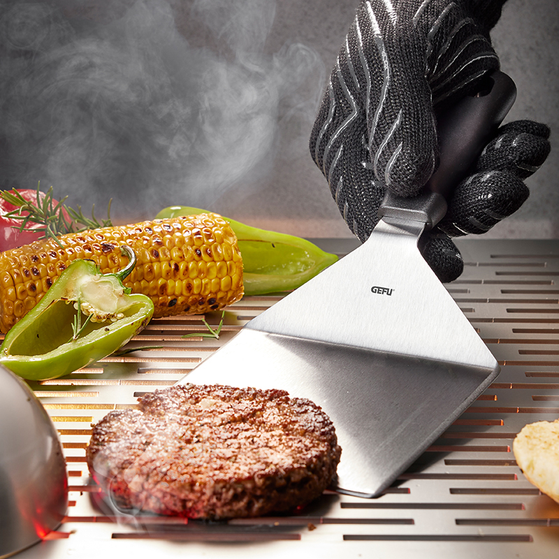 Profi-Grillwender beim Wenden eines saftigen Burgers auf dem Grill Ein Grillwender hält einen Burger auf einem Grill, begleitet von Maiskolben und grünem Paprika