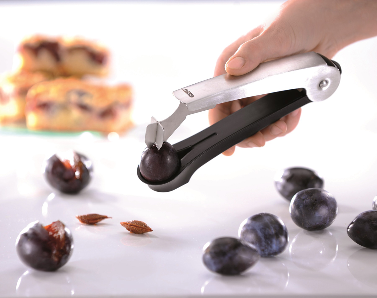 Praktischer Entkerner für frische Pflaumen Eine Hand verwendet einen Entkerner, um einen Pflaumenkern zu entfernen. Im Hintergrund sind entkernte Pflaumen und Kuchenstücke zu sehen.