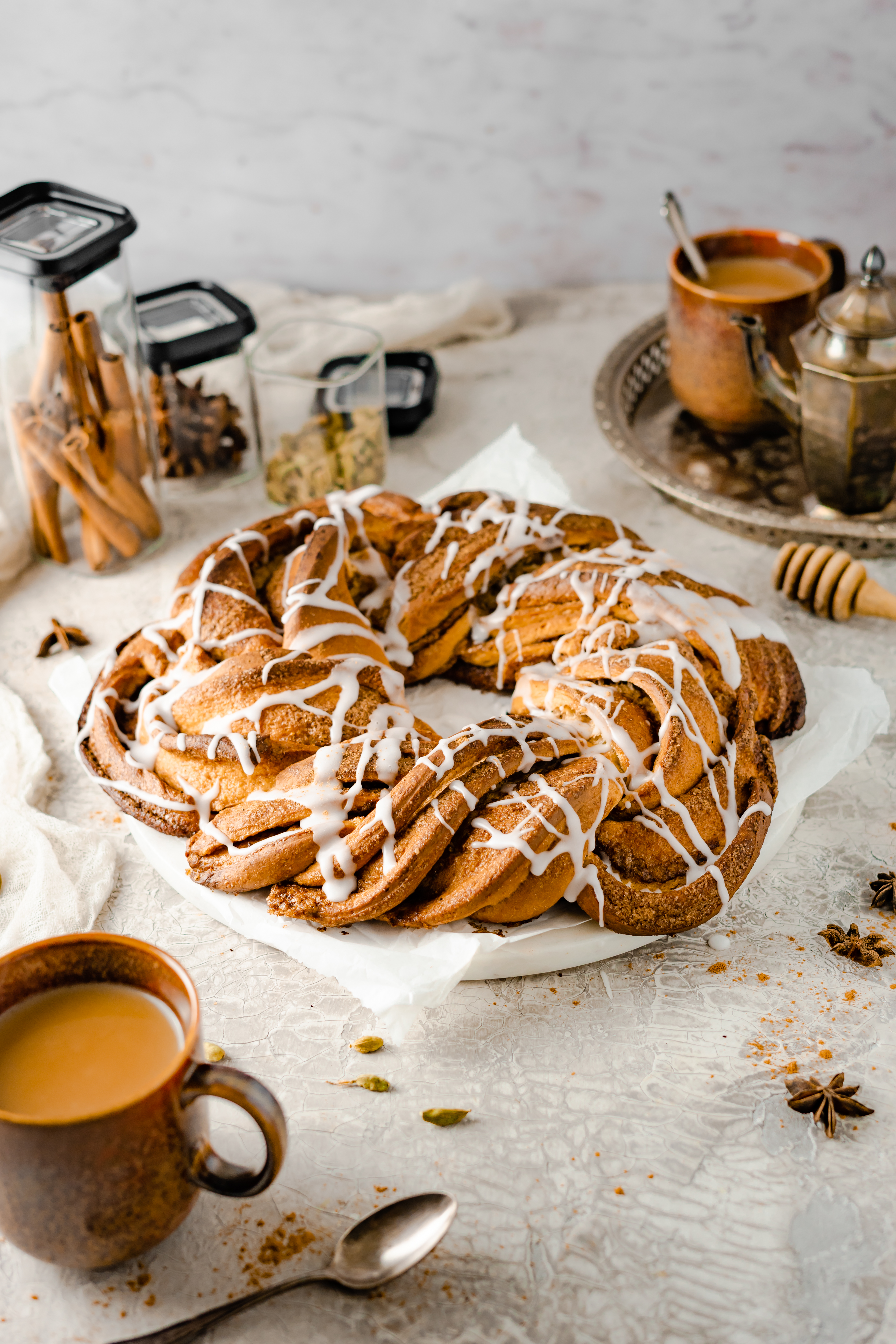 Chai Latte Hefezopf Große Zimtschnecke mit weißem Zuckerguss, umgeben von Gewürzgläsern, einer Kaffeetasse und einer Teekanne