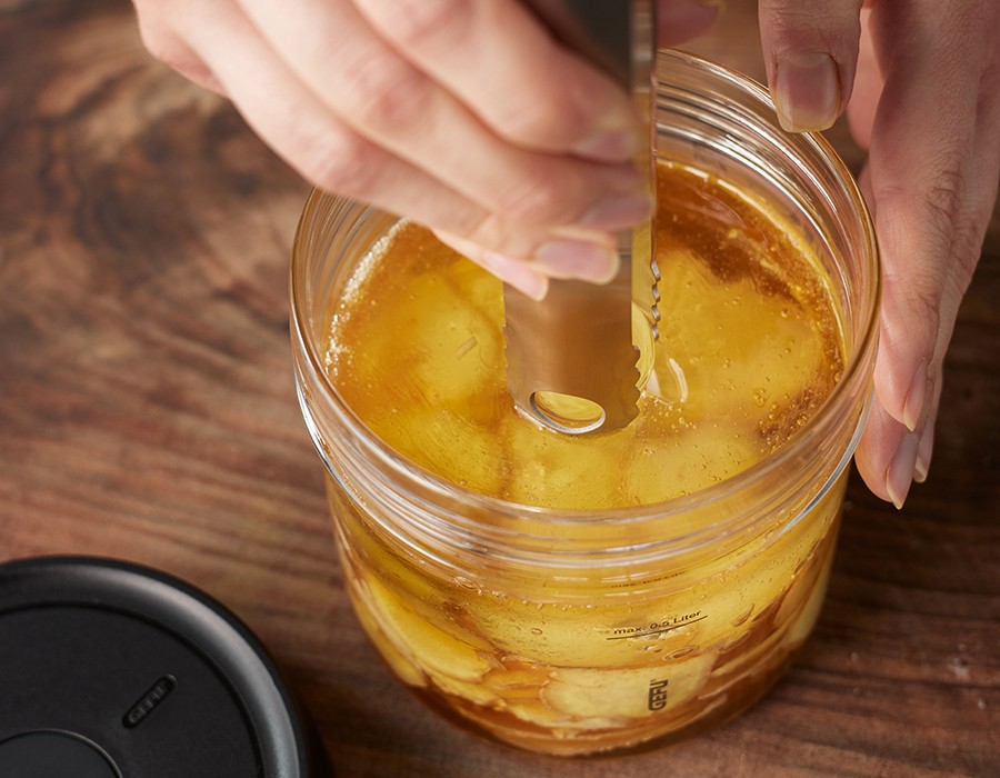 Fermentierter Knoblauch in Waldhonig Nahaufnahme einer Hand, die einen Gurkenzangenöffner in ein Glas mit eingelegten Gurken taucht.