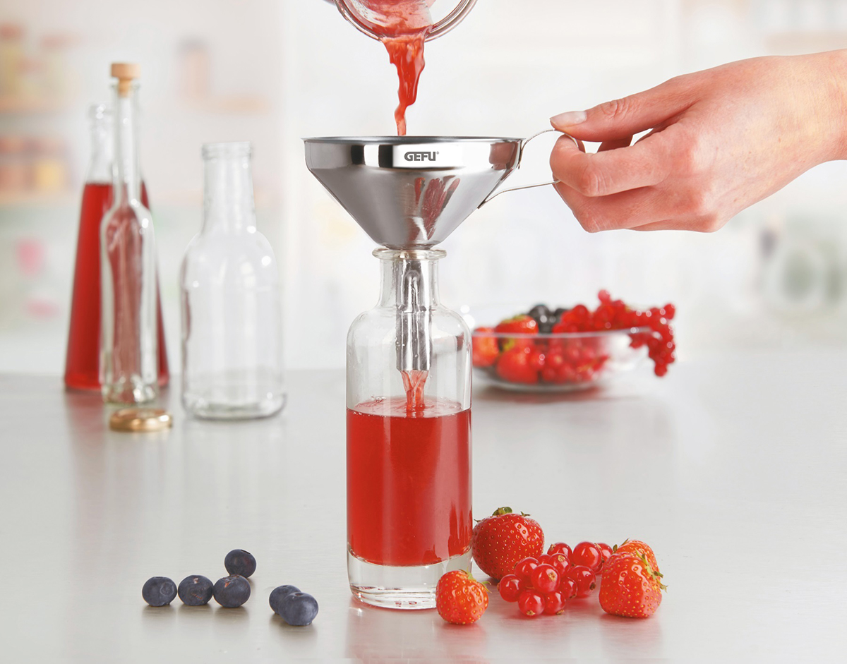 Praktischer Trichter zum Abfüllen von Fruchtsaft Ein Fruchtsaft wird mit einem Edelstahltrichter in eine Glasflasche abgefüllt, umgeben von Beeren auf einem Tisch.