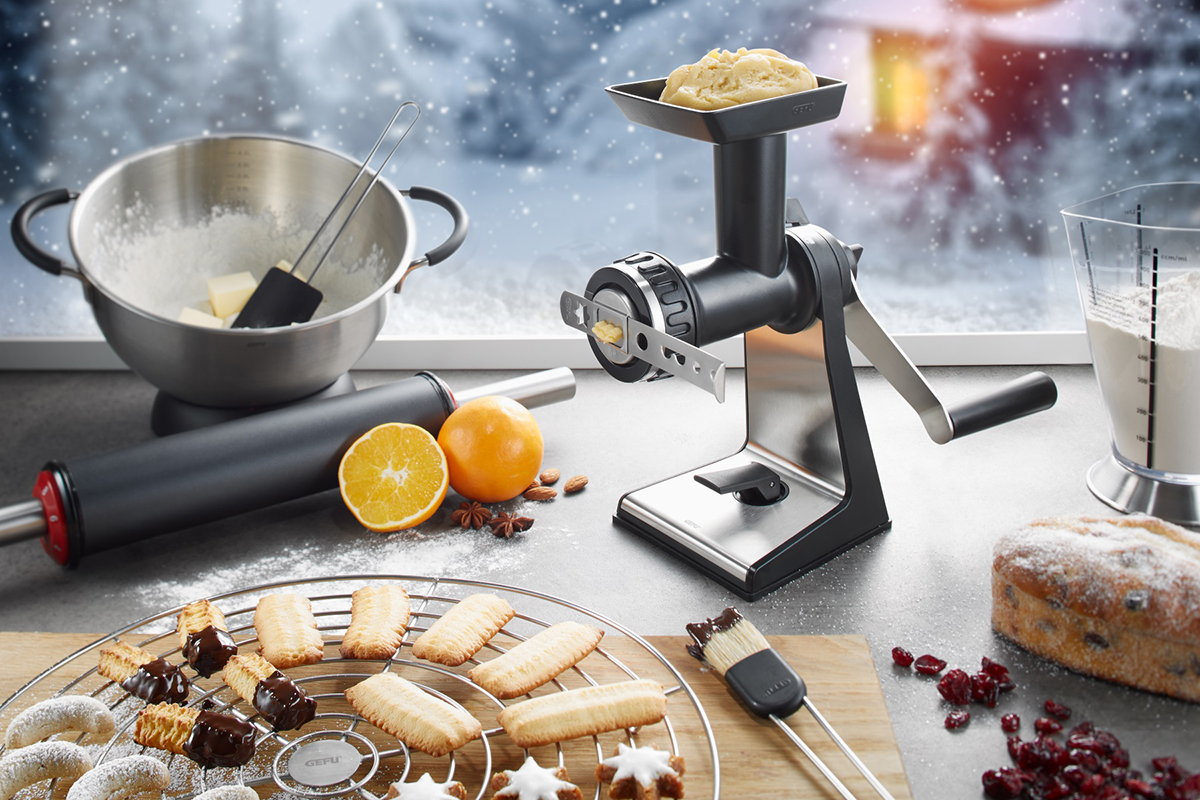 Weihnachtliche Kekszubereitung mit Gebäckpresse und frischen Zutaten Eine moderne Gebäckpresse auf einem Tisch, umgeben von Zutaten wie Mehl, Butter und Orangen, mit frisch gebackenen Keksen im Vordergrund, während draußen Schnee fällt.