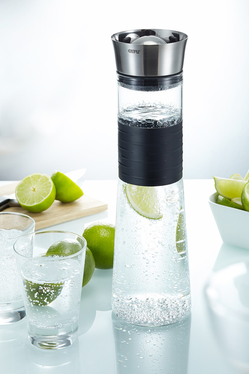 Stylische Glaskaraffe mit integriertem Edelstahlsieb Eine moderne Glaskaraffe mit einem Edelstahlaufsatz, gefüllt mit sprudelndem Wasser und Limettenscheiben, daneben Gläser und frische Limetten.
