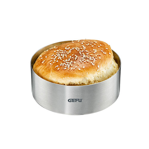 Ein runder Edelstahl-Brötchenformer der Marke GEFU, gefüllt mit einem frisch gebackenen Sesambrötchen, auf weißem Hintergrund.