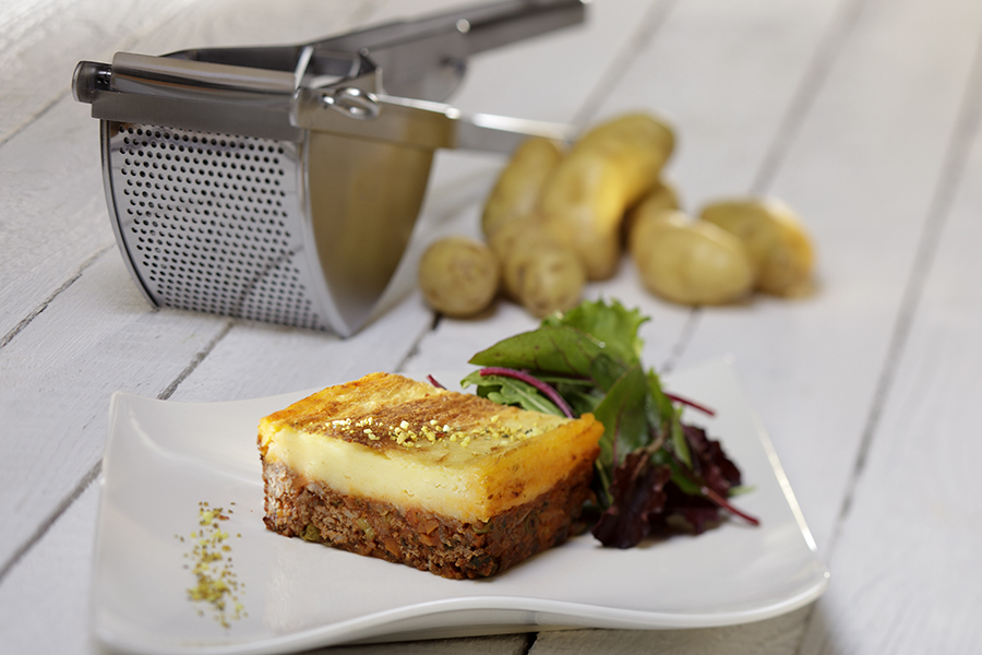 Cottage Pie Ein Stück Pastitsio auf einem weißen Teller, serviert mit grünem Salat. Im Hintergrund sind eine Kartoffelpresse und einige Kartoffeln zu sehen.