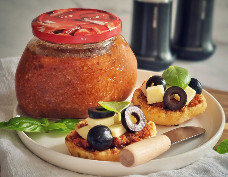 Aufstrich Ein Glas selbstgemachtes Pesto neben belegten Brötchen mit Käse, schwarzen Oliven und Basilikum auf einem Teller.
