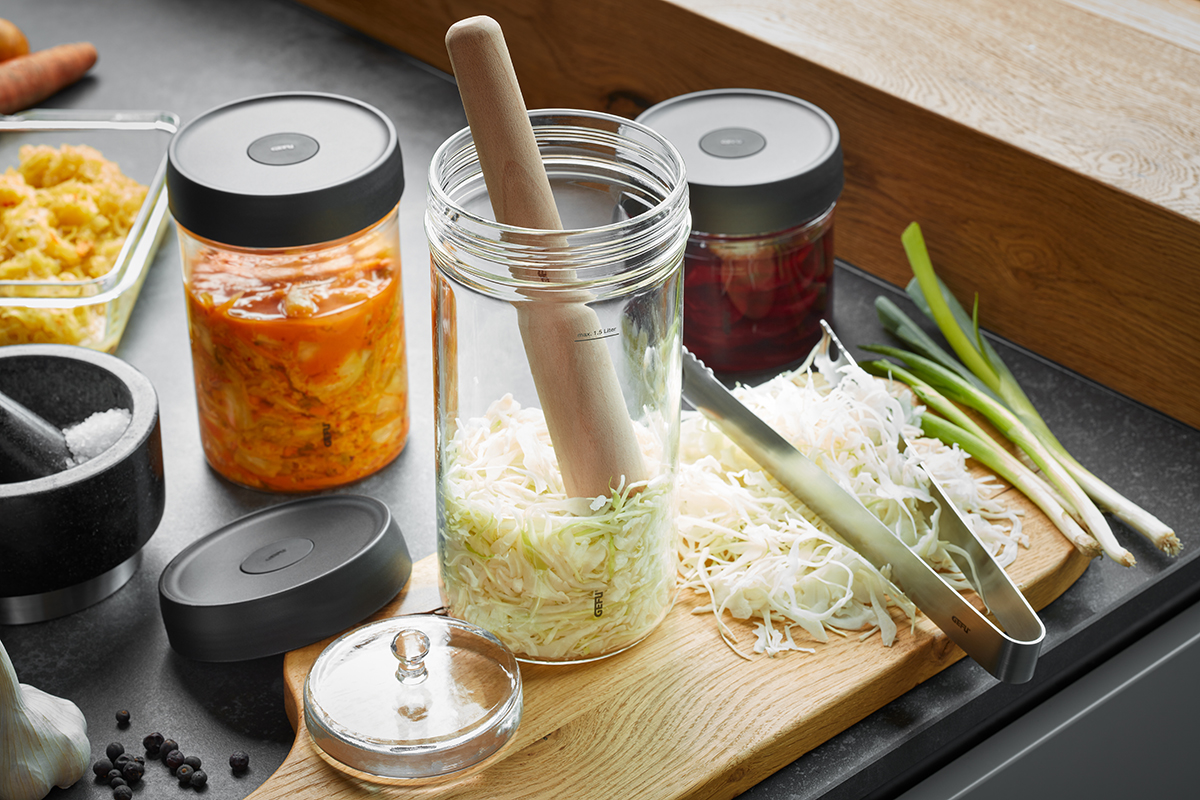 Fermentationsset für Sauerkraut und Gemüse mit Glasbehälter und Stampfer Bild eines Fermentationssets mit Glasbehälter, Stampfer und verschiedenen fermentierten Gemüsesorten auf einem Küchentisch.
