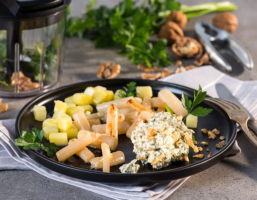 Petersilien-Walnuss-Dip mit gebratenen Schwarzwurzeln  Ein Teller mit gebratenem Spargel, gewürfelten Kartoffeln und einem Walnuss-Käse-Dip, umgeben von frischen Kräutern und Walnüssen, auf einer grauen Tischdecke. In der Nähe ein Kräutermixer und ein Nussknacker.
