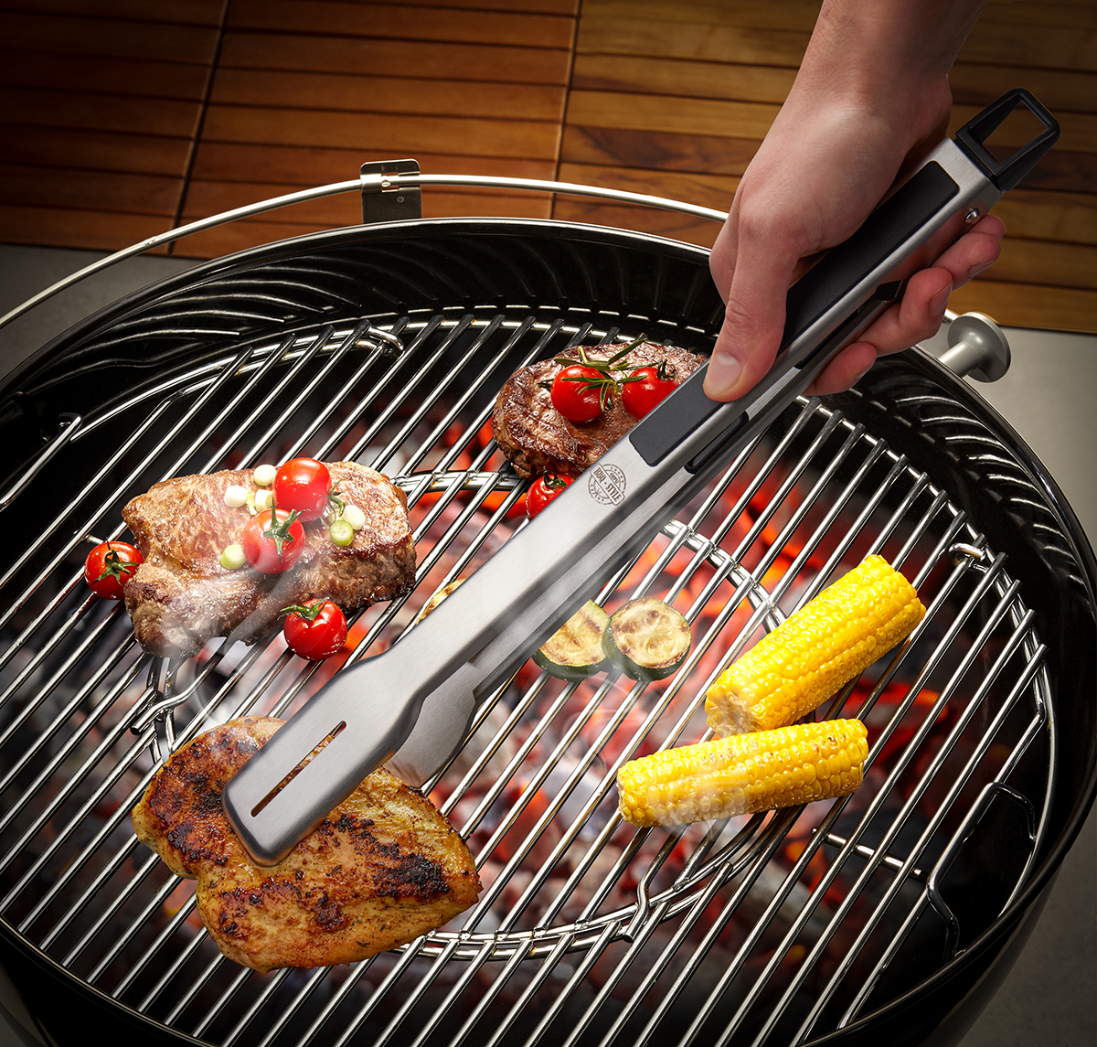 Eine Grillzange hält ein Steak auf einem Holzkohlegrill mit Gemüse und Maiskolben, umgeben von glühender Kohle.