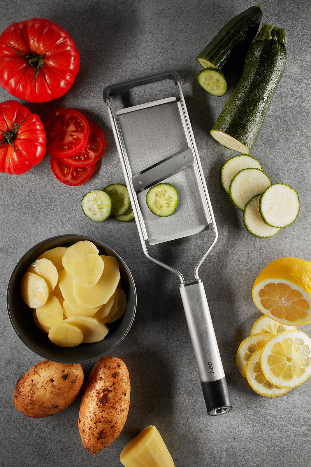 Gemüsehobel mit frisch geschnittenem Gemüse und Obst auf Küchentheke Gemüsehobel neben Tomaten, Kartoffeln, Gurken, Zucchini und Zitronenscheiben auf einer grauen Oberfläche