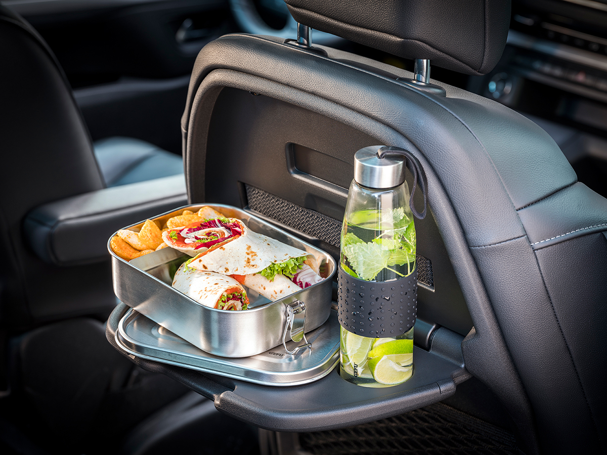 Praktische Lunchbox aus Edelstahl und Trinkflasche im Auto Eine Lunchbox aus Edelstahl mit Wraps und Chips neben einer Trinkflasche mit Limetten und Minze auf einem klappbaren Autositztablett.