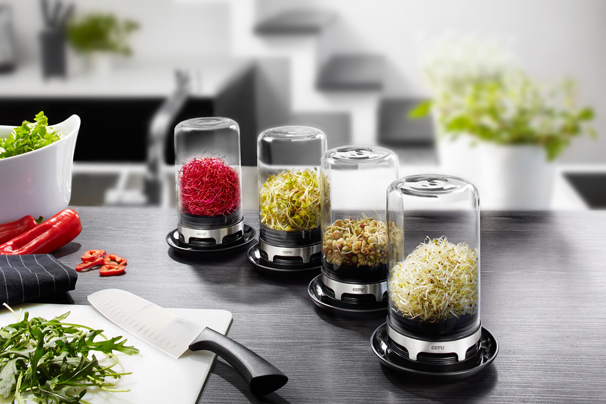 Frische Sprossen und Keimlinge in modernen Sprout Containern Verschiedene Sprout Container mit frischen Sprossen und Keimlingen auf einem Küchentisch, umgeben von frischem Gemüse und einem Messer.