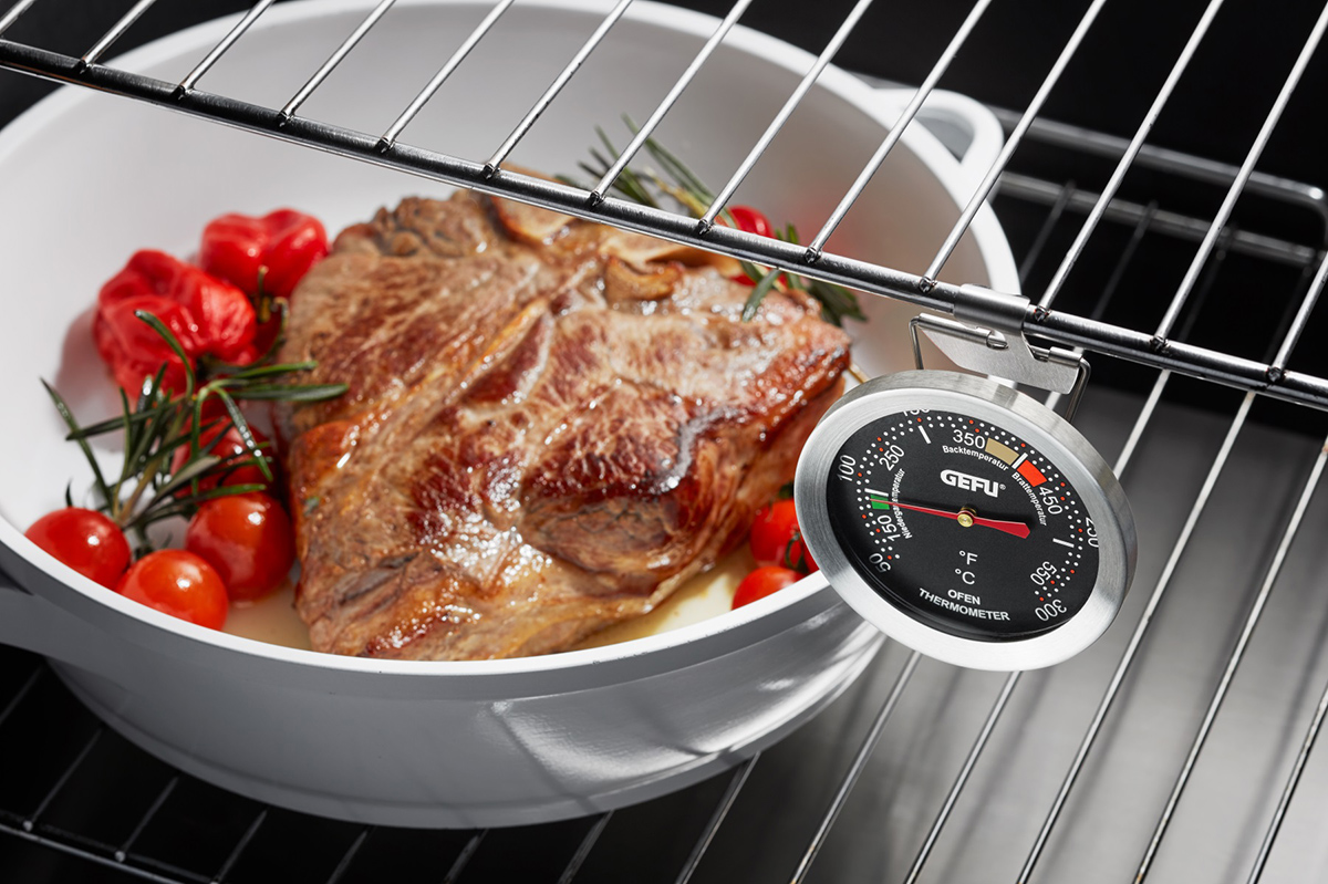 Backofen-Thermometer im Einsatz mit Bratenthermometer und Gemüse Ein Backofen-Thermometer neben einem Braten mit Tomaten und Rosmarin in einer weißen Bratpfanne im Ofen.