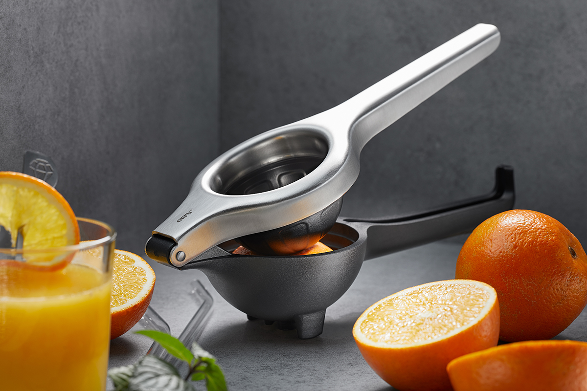 Manuelle Entsafterpresse aus Metall mit frischen Orangen Manuelle Entsafterpresse aus Edelstahl mit frischen Orangen und einem Glas Orangensaft auf einem Tisch