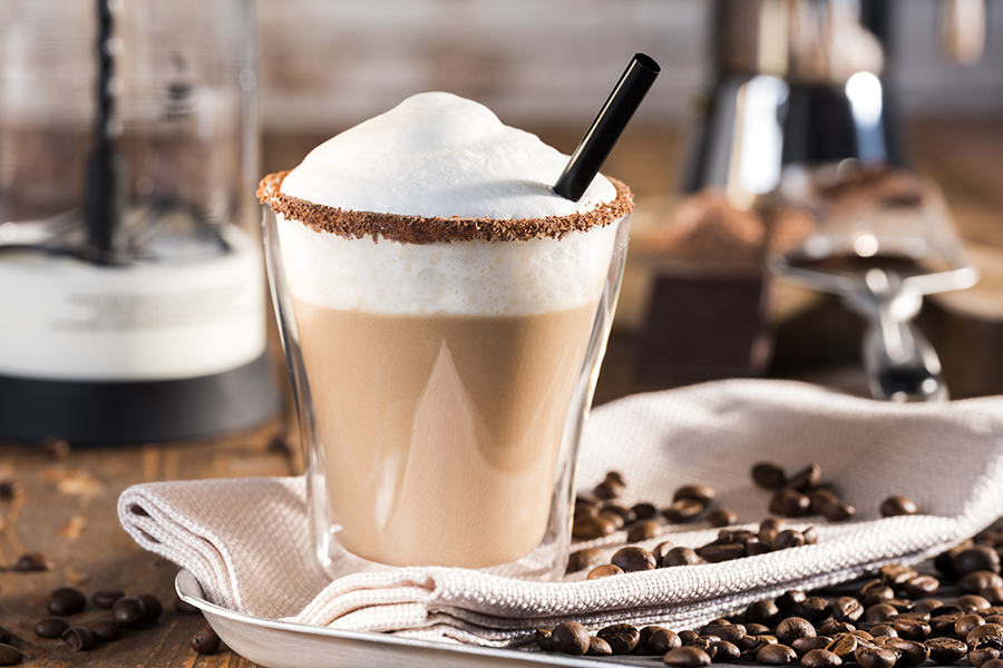 Caffè con Yoghurt Ein Glas Cappuccino mit cremiger Sahnehaube und Schokoladenrand, umgeben von Kaffeebohnen auf einem Tuch
