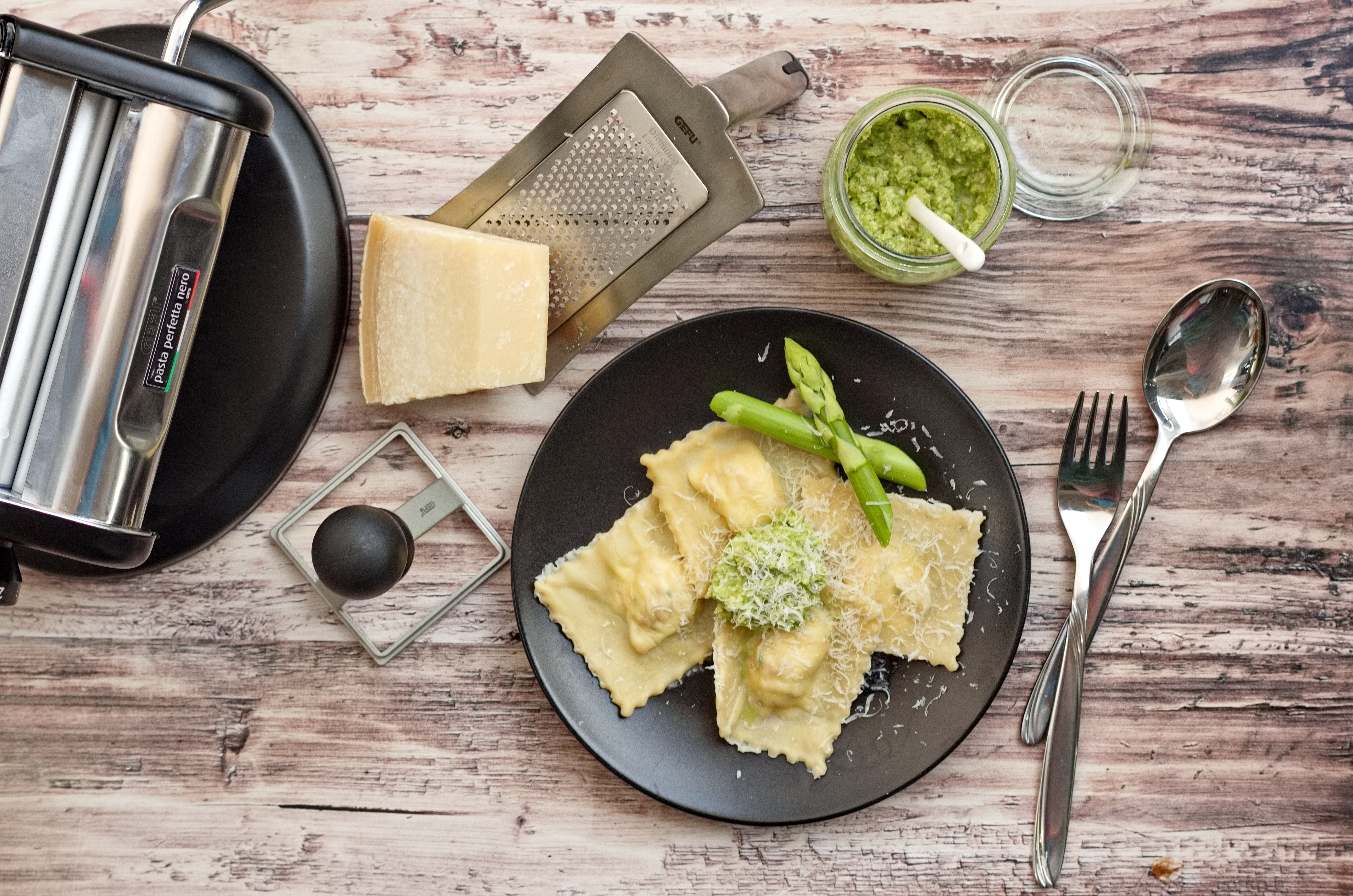 Spargel Ravioli Ein Teller mit selbstgemachten Ravioli, garniert mit grünem Spargel und Parmesan, daneben eine Pasta-Maschine und geriebener Käse.