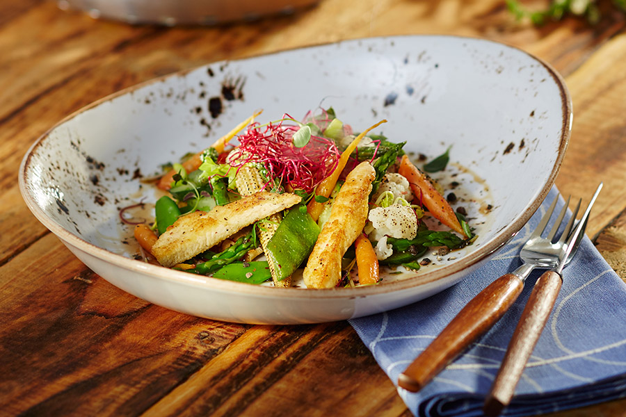 Asiagemüse / Hähnchenbrust-Streifen Ein Teller mit frischem Gemüsesalat und gebratenem Halloumi, serviert auf einem Holztisch mit Besteck auf einer blauen Serviette