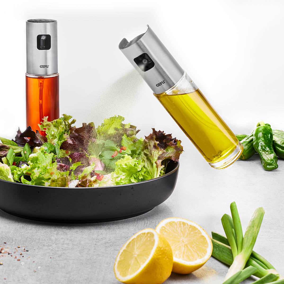 Zwei Sprüher mit Öl und Essig neben einem knackigen Salat in einer schwarzen Schüssel, umgeben von Zitronenhälften und Frühlingszwiebeln.