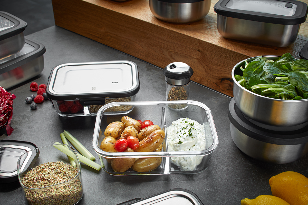 Moderne Küchenutensilien aus Edelstahl und Glas mit Lebensmitteln Verschiedene Küchenbehälter aus Glas und Edelstahl, gefüllt mit Salat, Kartoffeln, Tomaten und Kräuterdip, auf einem Tisch mit Beeren und Zitronen.