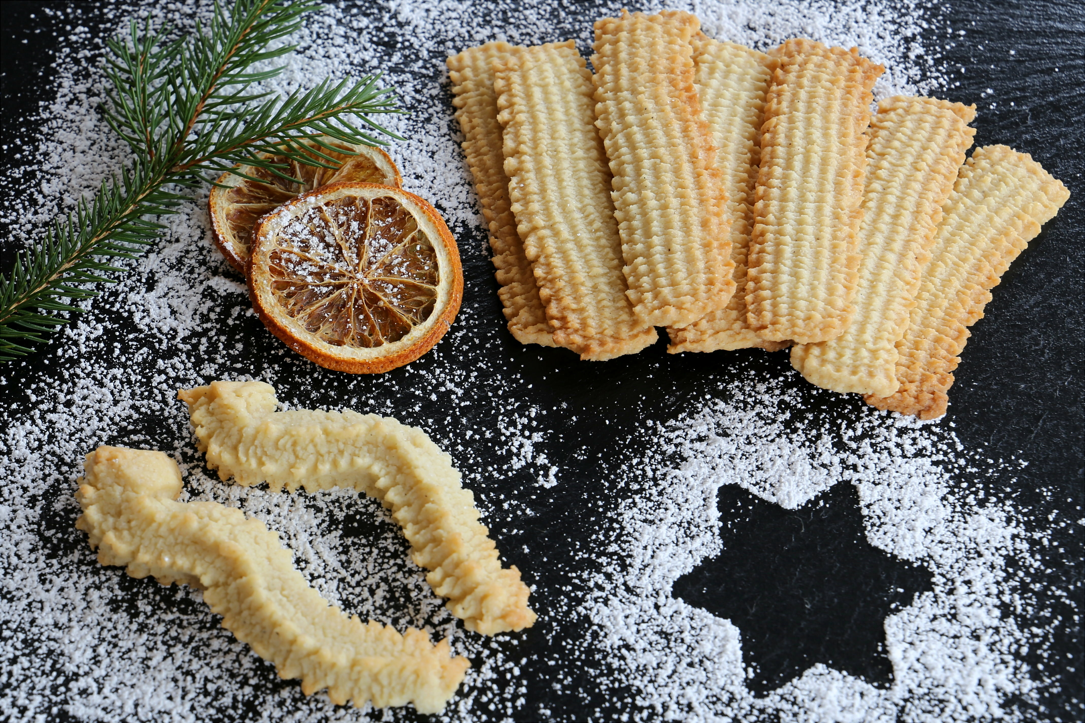Omas Spritzgebäck Knuspriges Spritzgebäck mit getrockneten Orangenscheiben, Tannenzweig und Puderzucker auf dunkler Fläche