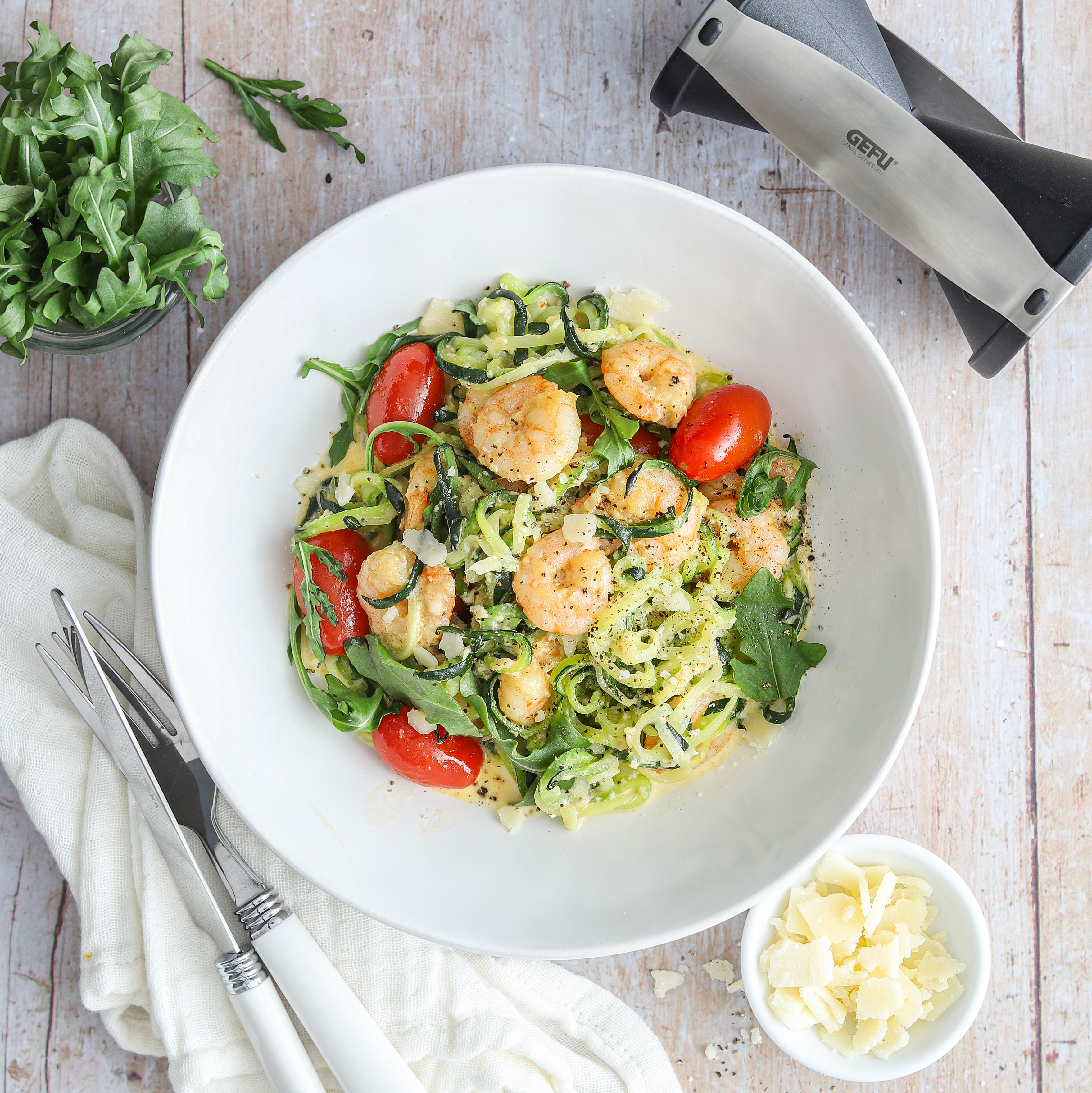 Zoodles with shrimps in cream sauce Ein Teller mit Zucchini-Nudeln, garniert mit Garnelen, Kirschtomaten, Rucola und Käse, daneben Besteck und eine Schüssel mit Parmesanspänen.
