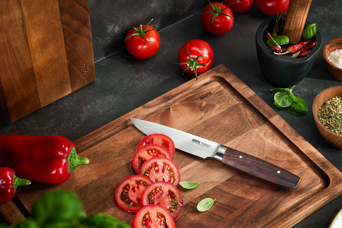 Hochwertiges Küchenmesser auf einem Holzschneidebrett mit frischen Tomaten und Gewürzen Ein Küchenmesser liegt auf einem Holzschneidebrett, umgeben von geschnittenen Tomaten, ganzen Tomaten, einer roten Paprika und Gewürzen.