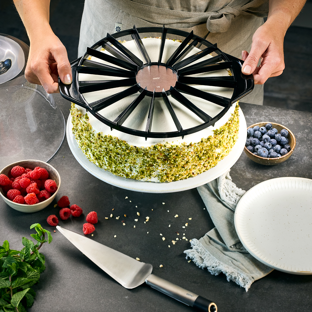 Eine Person portioniert einen mit Pistazien dekorierten Kuchen mithilfe eines Tortenschneiders, umgeben von frischen Himbeeren und Blaubeeren.