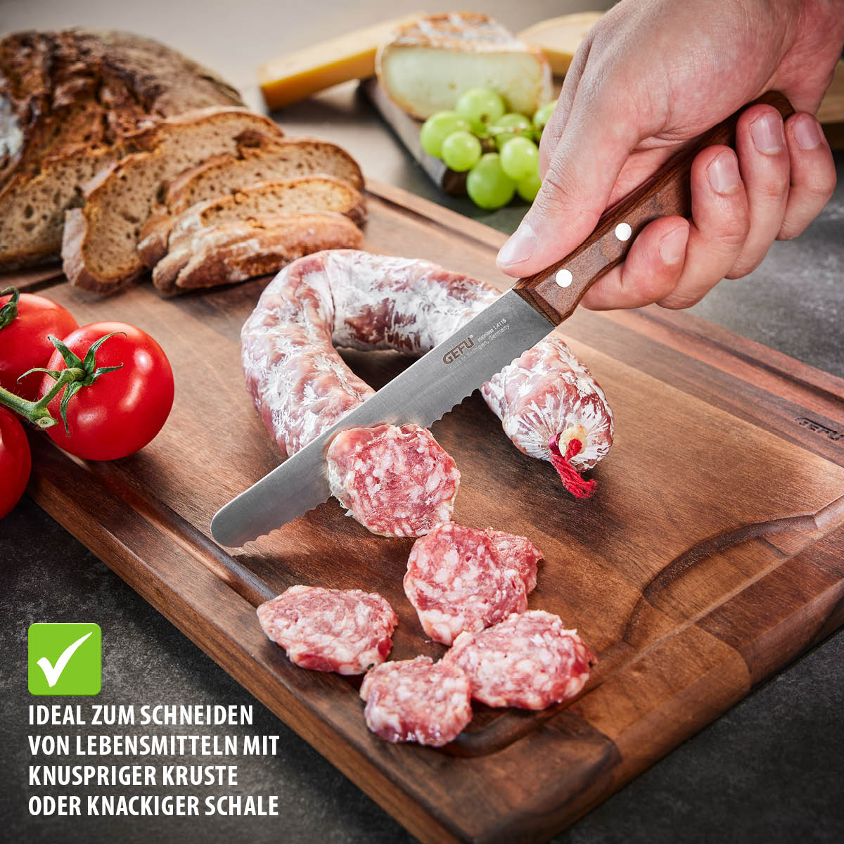 Schneidemesser auf Holzbrett beim Schneiden einer Salami, umgeben von Brot, Tomaten und Trauben