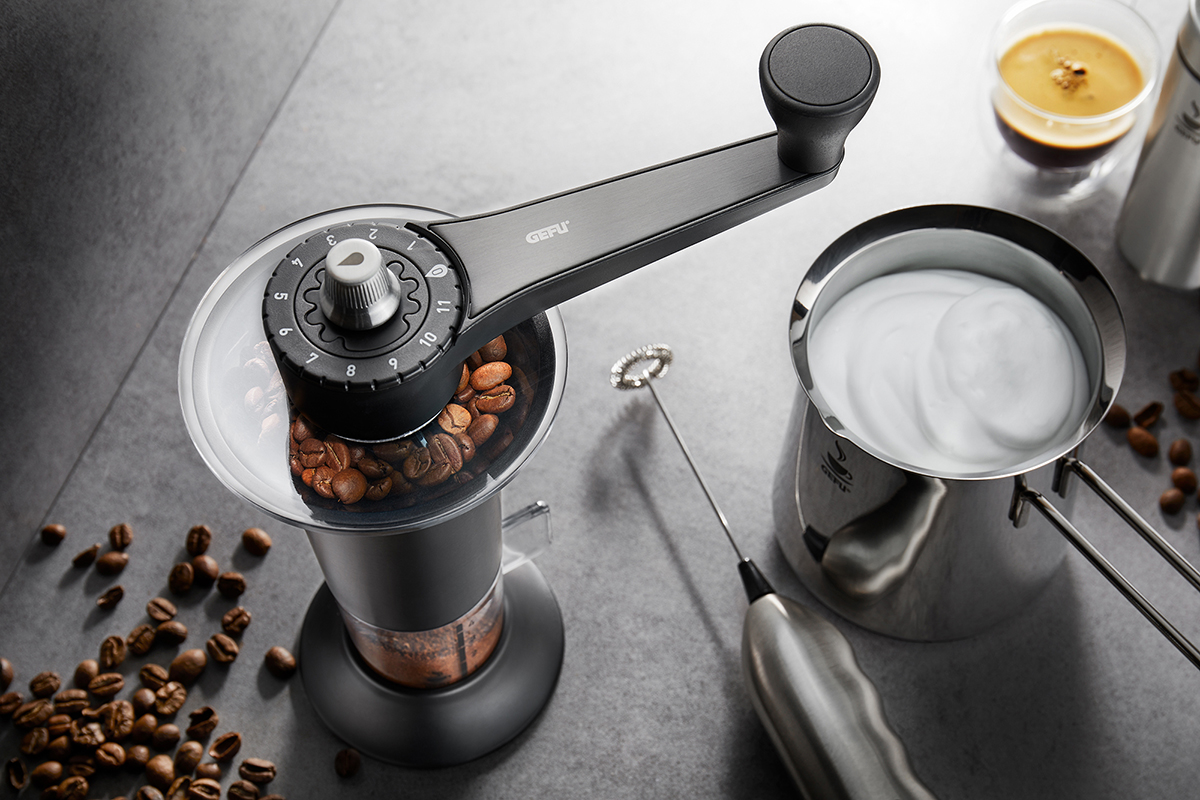 Nahaufnahme einer manuellen Kaffeemühle, umgeben von Kaffeebohnen und einem Milchaufschäumer neben einer Tasse Cappuccino