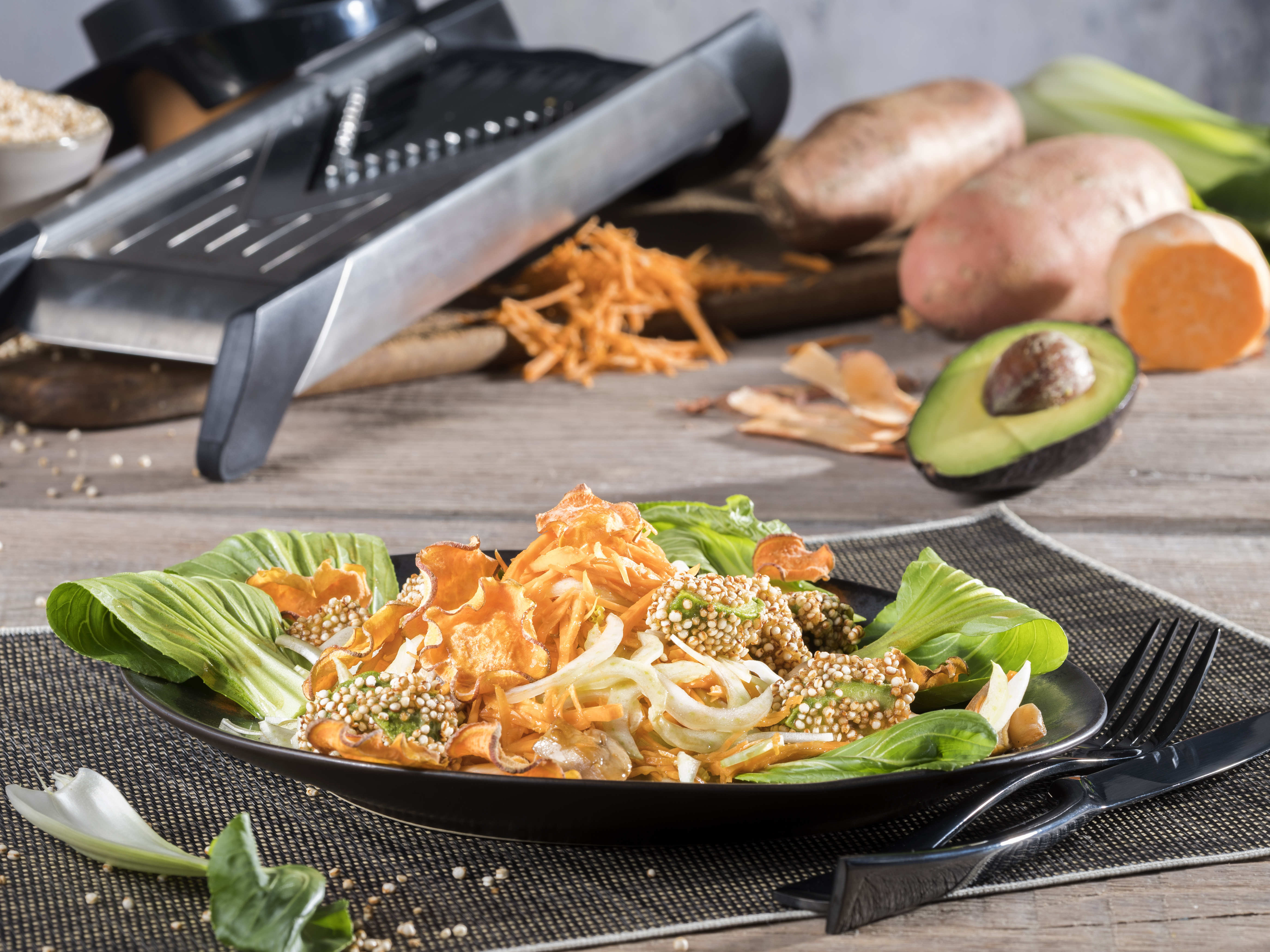 Süßkartoffelsalat mit Pak Choi, knusprigen Avocado und Süßkartoffelchips Ein Teller mit frischem Salat, Avocado und knusprigen Süßkartoffelchips vor einem Gemüseschneider und Süßkartoffeln im Hintergrund auf einem Holztisch