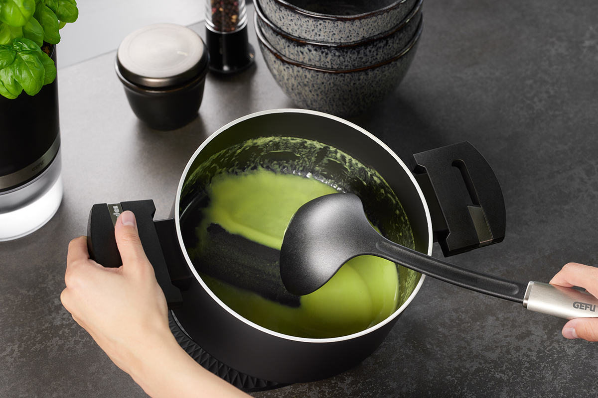 Kochtopf mit grünem Inhalt und schwarzem Schöpflöffel in moderner Küche Person hält einen Kochtopf mit grünem Inhalt, daneben ein schwarzer Schöpflöffel und Küchenutensilien auf einer Arbeitsfläche