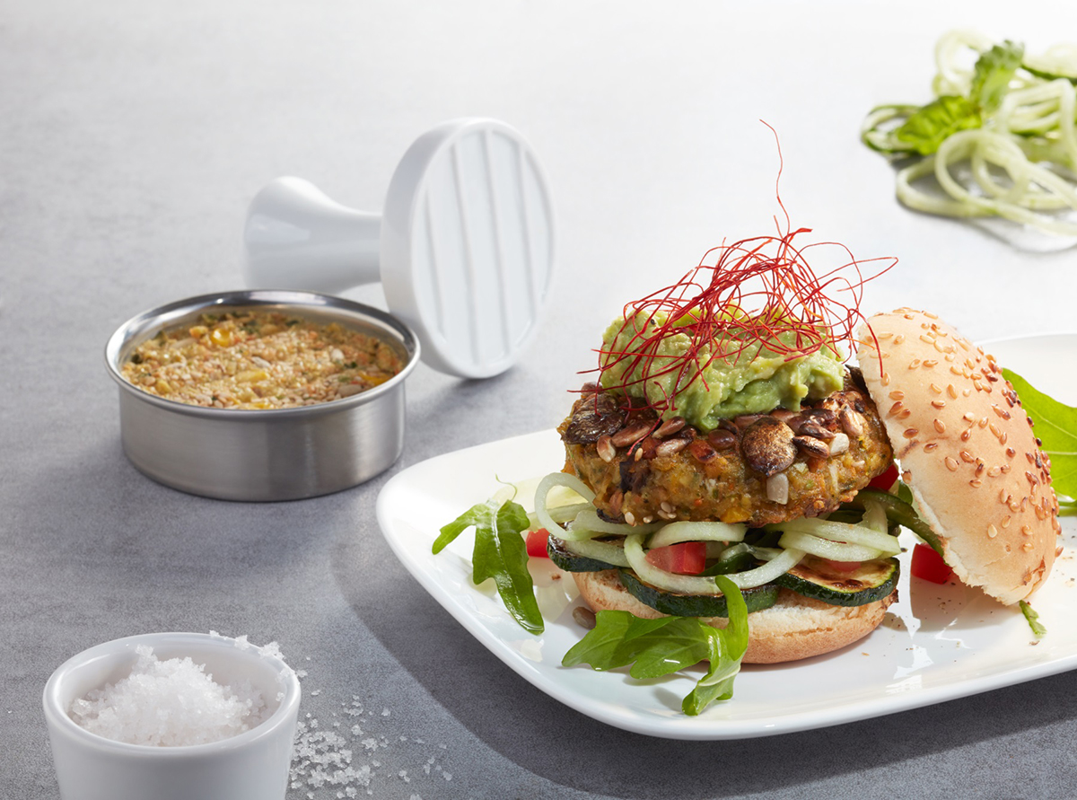 Leckerer vegetarischer Burger mit Gemüsepatty und Avocado Ein vegetarischer Burger mit Gemüsepatty, Avocado und frischem Salat, daneben eine Dose mit Zutaten und Salz auf einem Tisch.