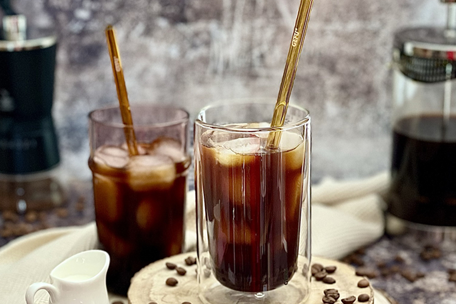 Cold Brew Cofffee Zwei Gläser mit Eiskaffee und Strohhalmen, umgeben von Kaffeebohnen und einer French Press auf einem Holztisch.