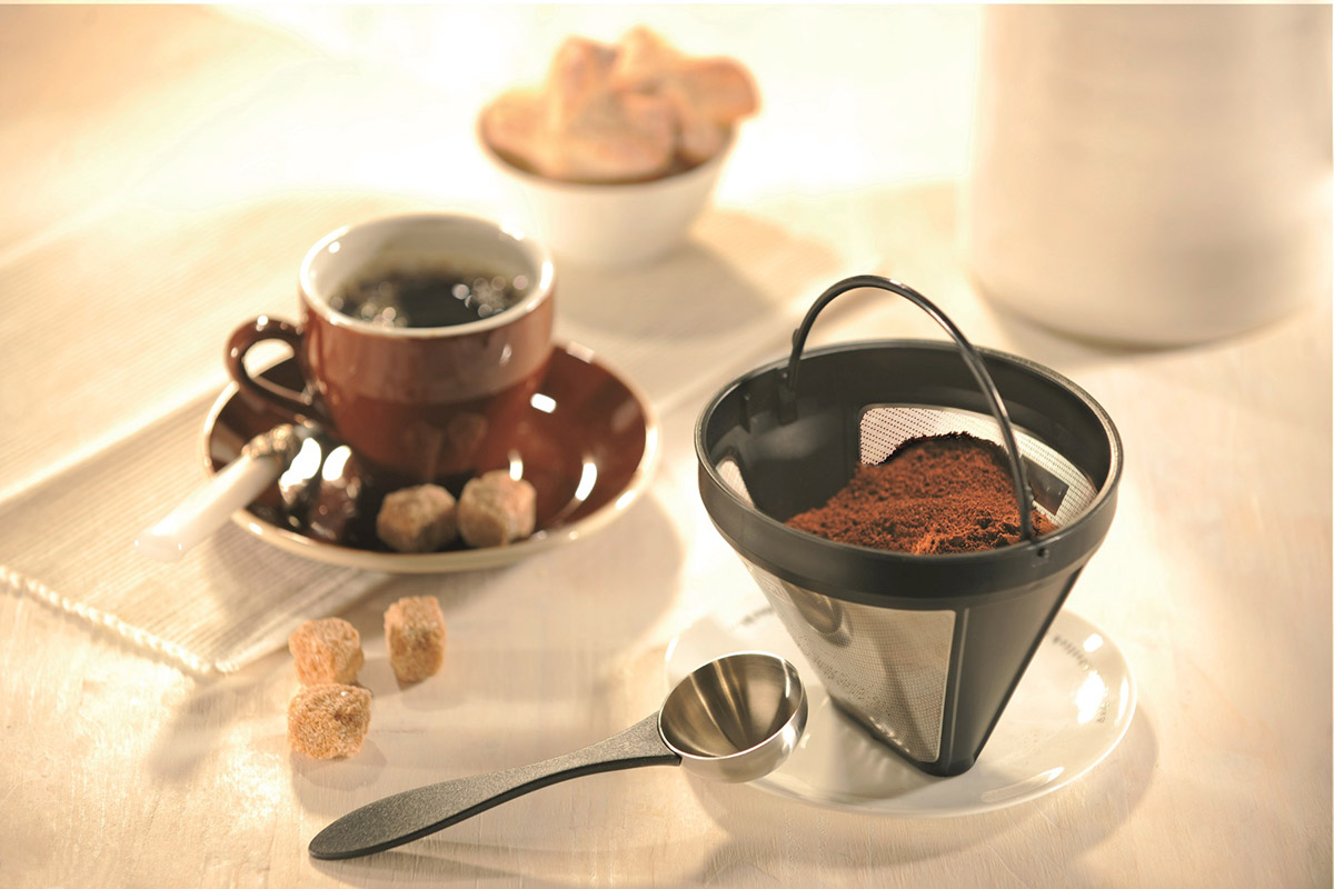 Kaffee-Set mit Filter und Tasse auf Tisch Eine Tasse Kaffee, ein Edelstahl-Kaffeefilter mit gemahlenem Kaffee und ein Löffel auf einem Tisch arrangiert.