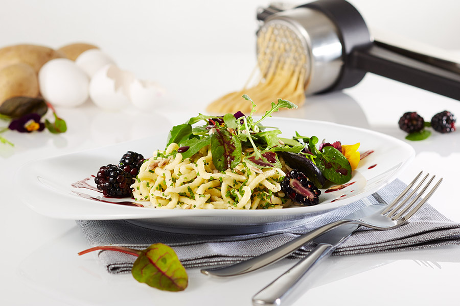 Butterspätzle mit Wildkräutersalat und Brombeerdressing Ein Teller mit Spätzle, garniert mit frischen Kräutern, Salat und Brombeeren, im Hintergrund eine Kartoffelpresse