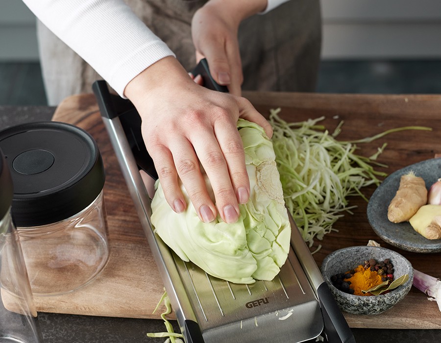 Koreanisches Kimchi Hand, die einen Kohlkopf auf einer Küchenreibe reibt, um Krautsalat zuzubereiten. Im Hintergrund sind Gewürze und ein Glas zu sehen.