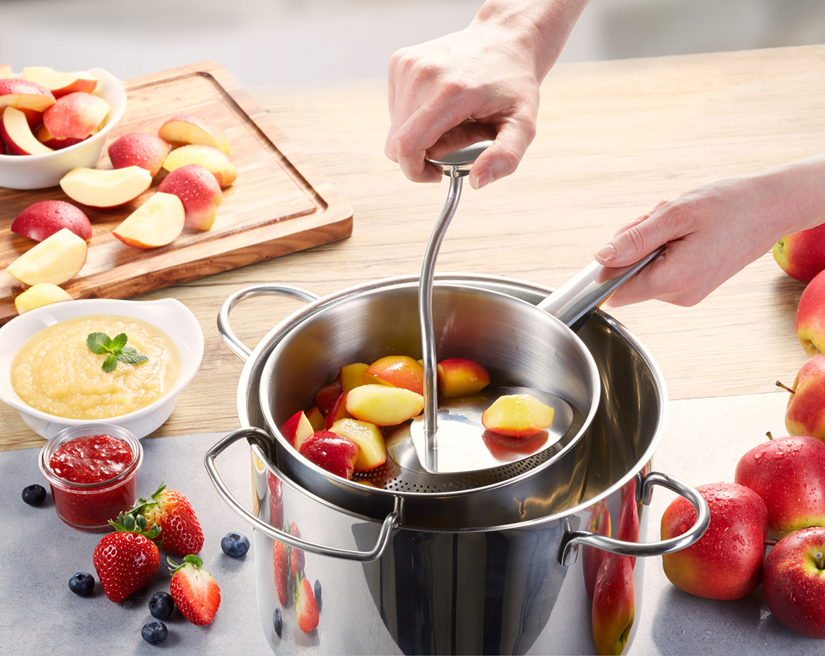Küchenhelfer: Passiermühle für frische Apfelmus-Zubereitung Person benutzt eine Passiermühle, um gekochte Äpfel in einem Topf auf einem Tisch mit geschnittenen Äpfeln, Apfelmus, Erdbeer- und Blaubeeren zu verarbeiten.