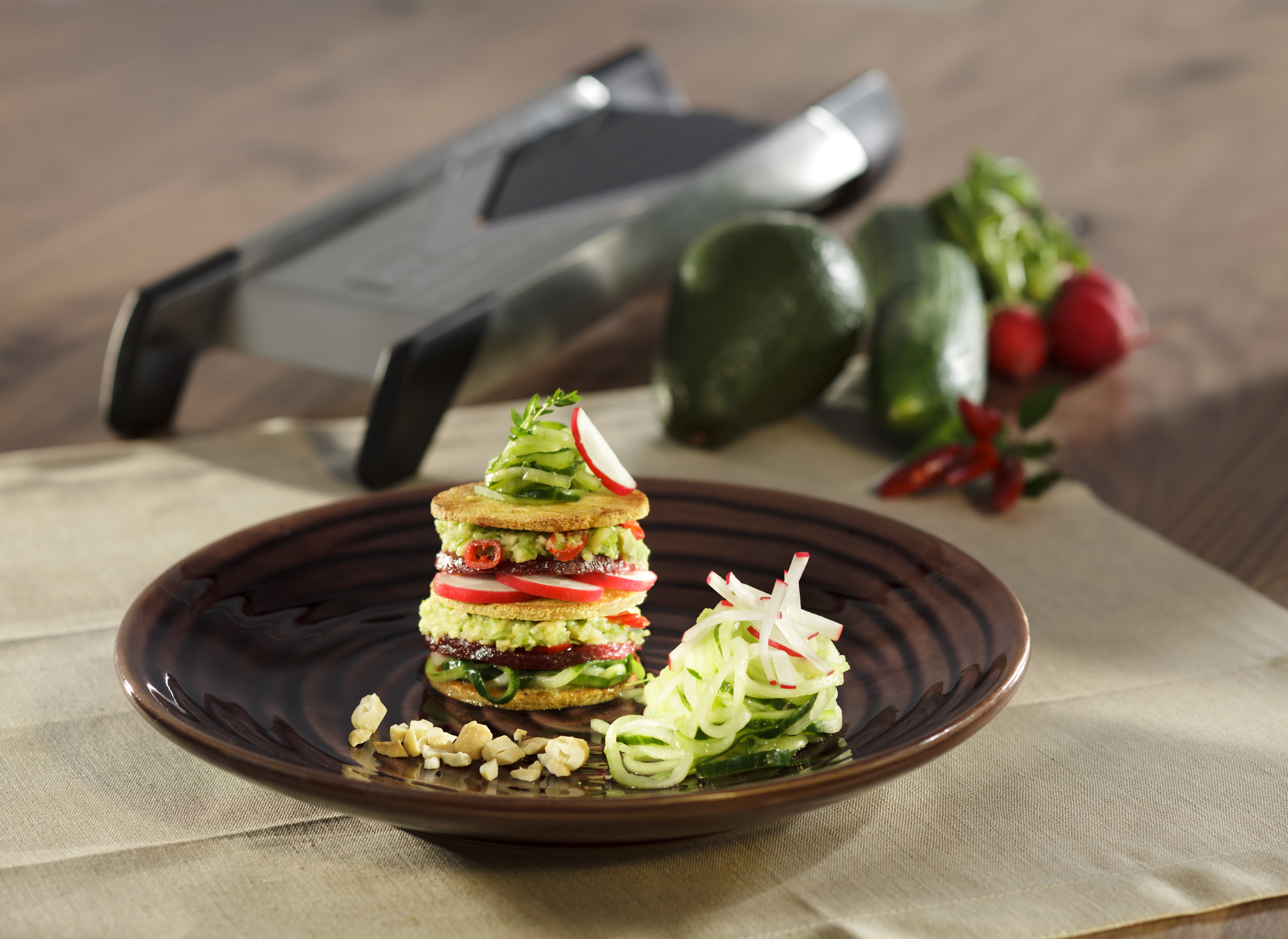 Tofu-Avocado Törtchen mit japanischem Gurkensalat Ein kunstvoll angerichteter Gemüse-Turm auf braunem Teller mit frischen Gemüsezutaten und einem Gemüseschneider im Hintergrund