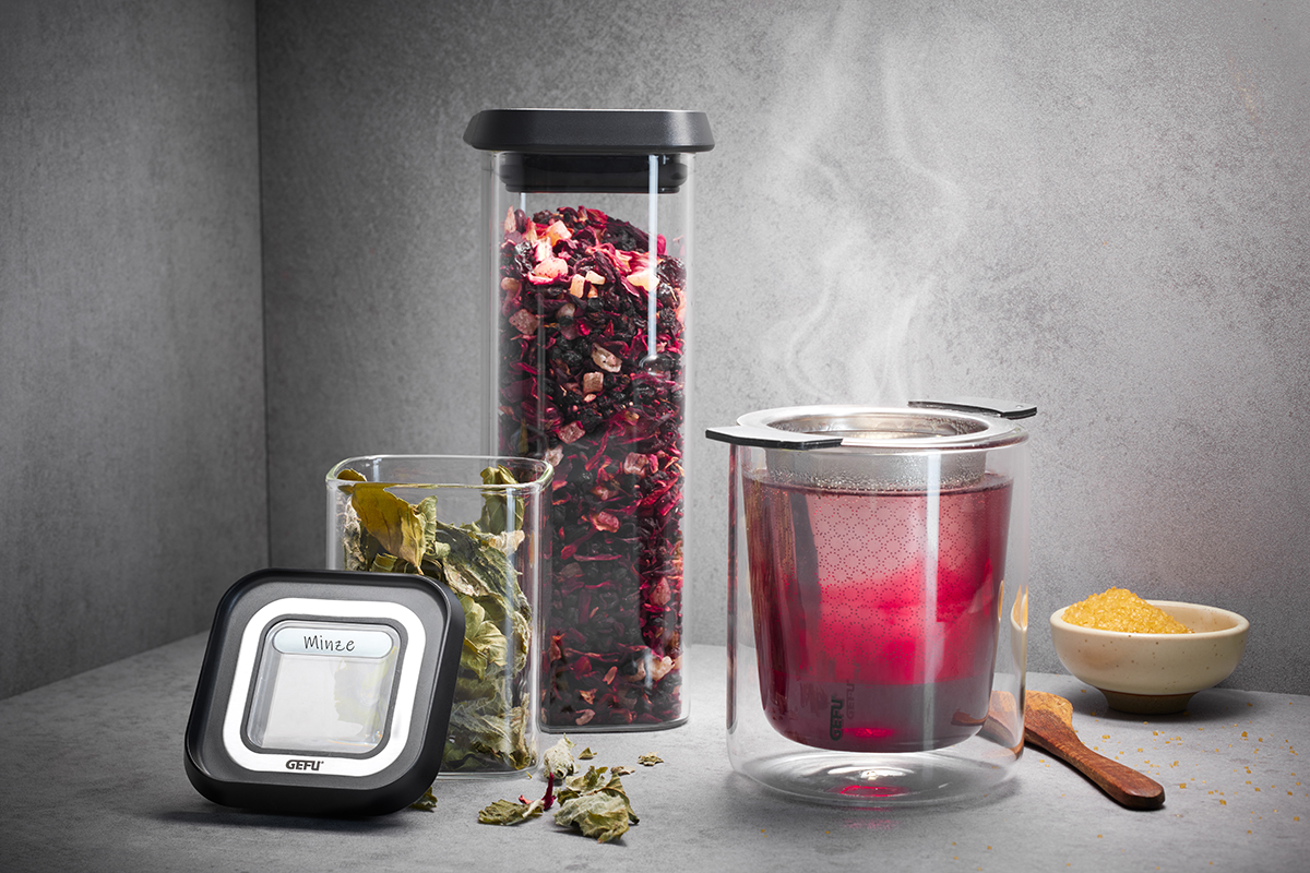 Set zur Teezubereitung mit Teeglas, Teesieb und Teebehältern Ein Set zur Teezubereitung mit einem doppelwandigen Teeglas, Teesieb, zwei verschiedenen Teebehältern und einer kleinen Schale Zucker auf grauem Hintergrund.