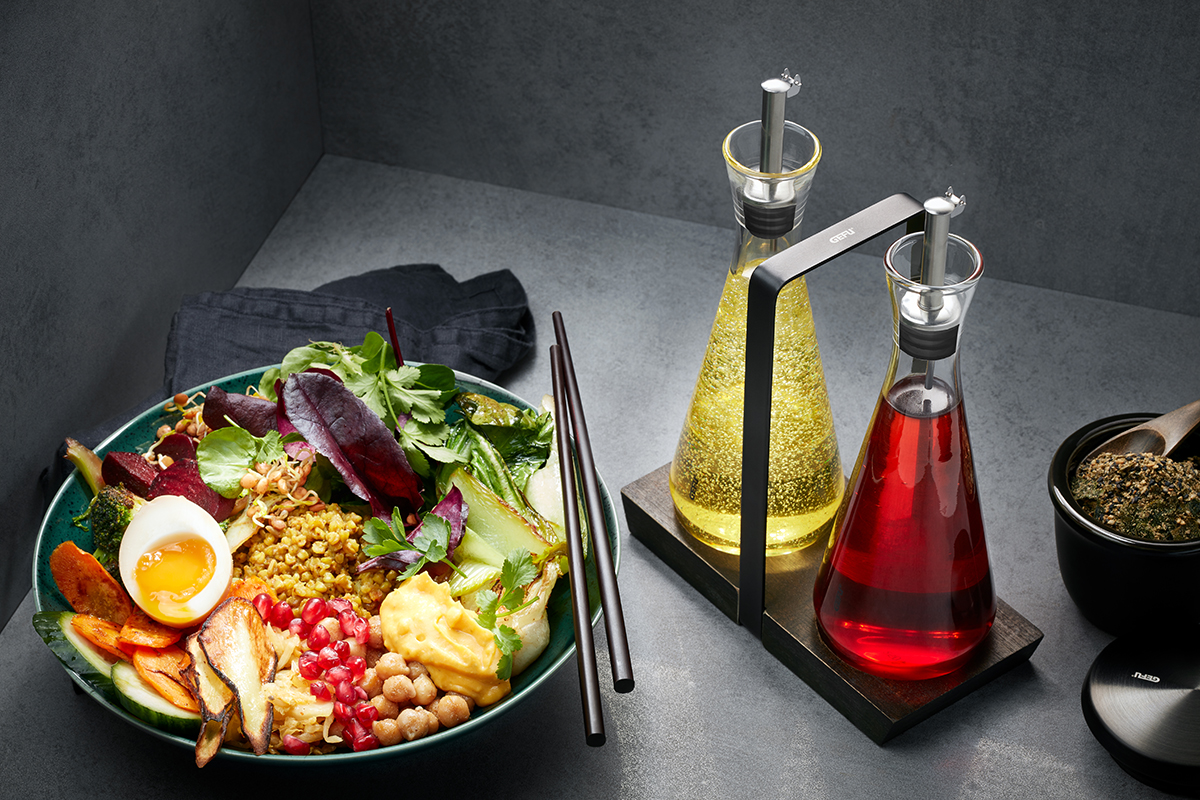 Gesunde Buddha-Bowl mit frischem Gemüse und Dressing-Set Eine Schüssel mit einer bunten Buddha-Bowl aus frischem Gemüse, einem Ei und Kräutern. Daneben steht ein Öl- und Essig-Set in modernen Karaffen.