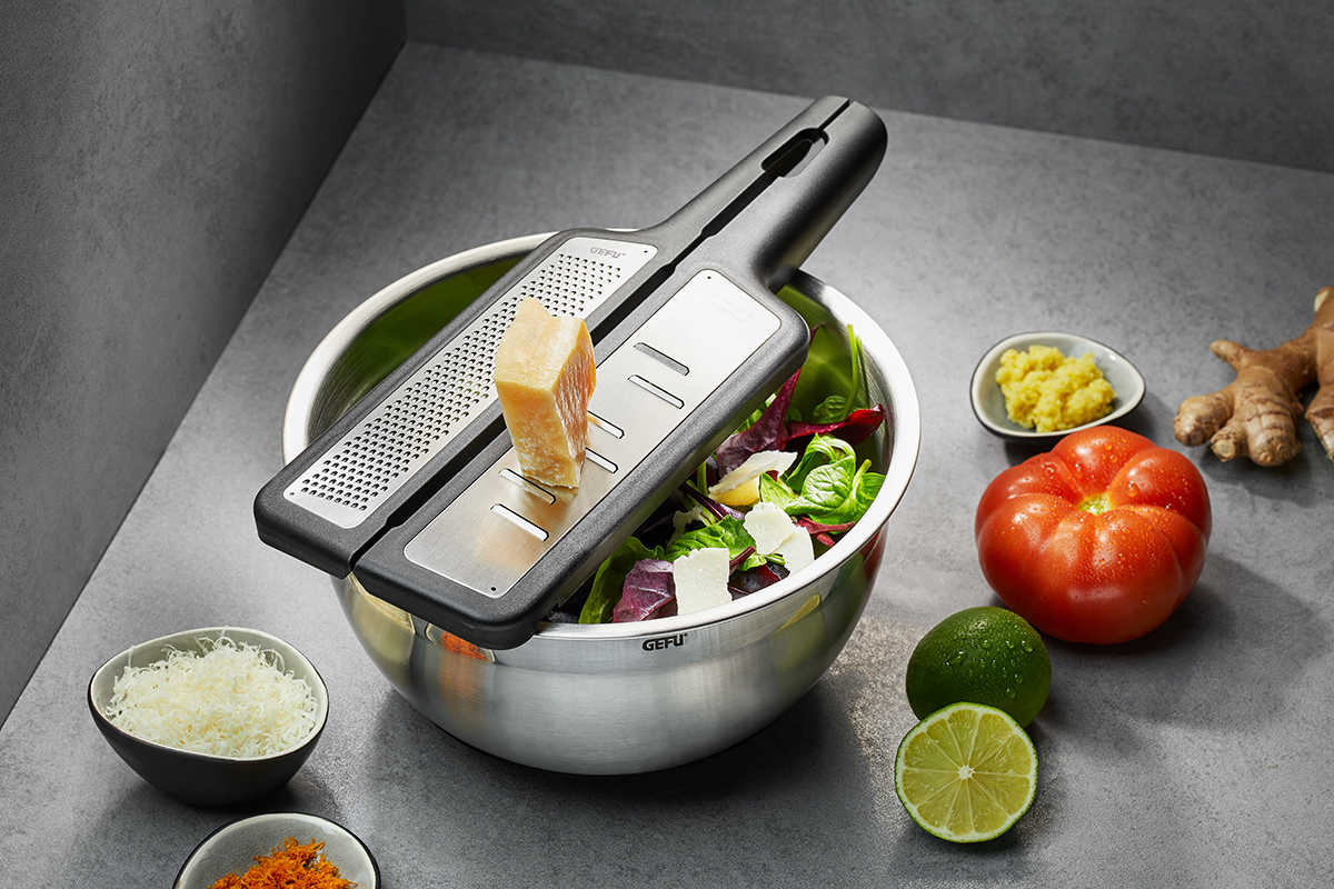 Multifunktionale Gemüsereibe über Salatschüssel für frische Zubereitung Gemüsereibe auf Edelstahlschüssel mit Parmesanstück, umgeben von Limette, Tomate und Ingwer