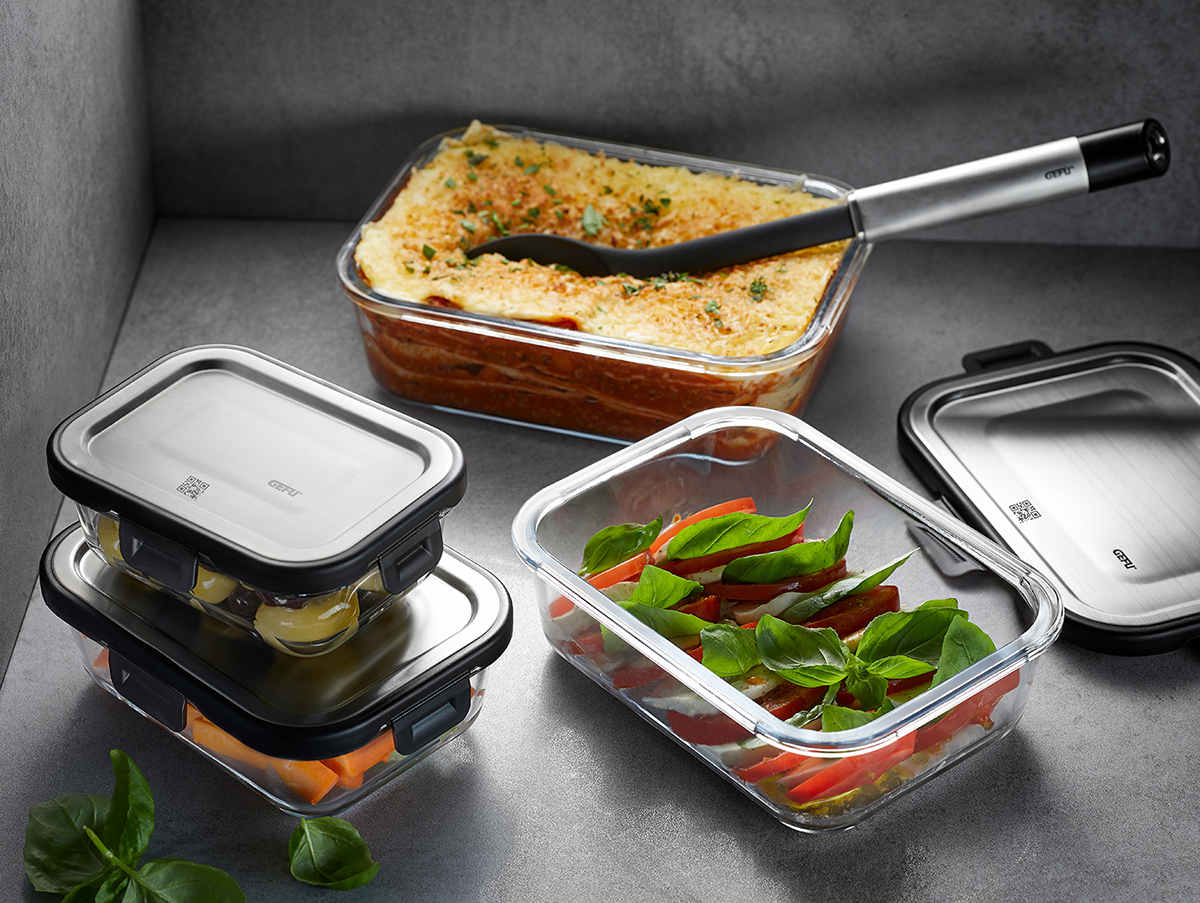 Glas-Behälter-Set mit Edelstahl-Deckel und frischen Zutaten Mehrere Glasbehälter mit Edelstahl-Deckeln, gefüllt mit Lasagne, Salat und Gemüse, auf einem grauen Untergrund