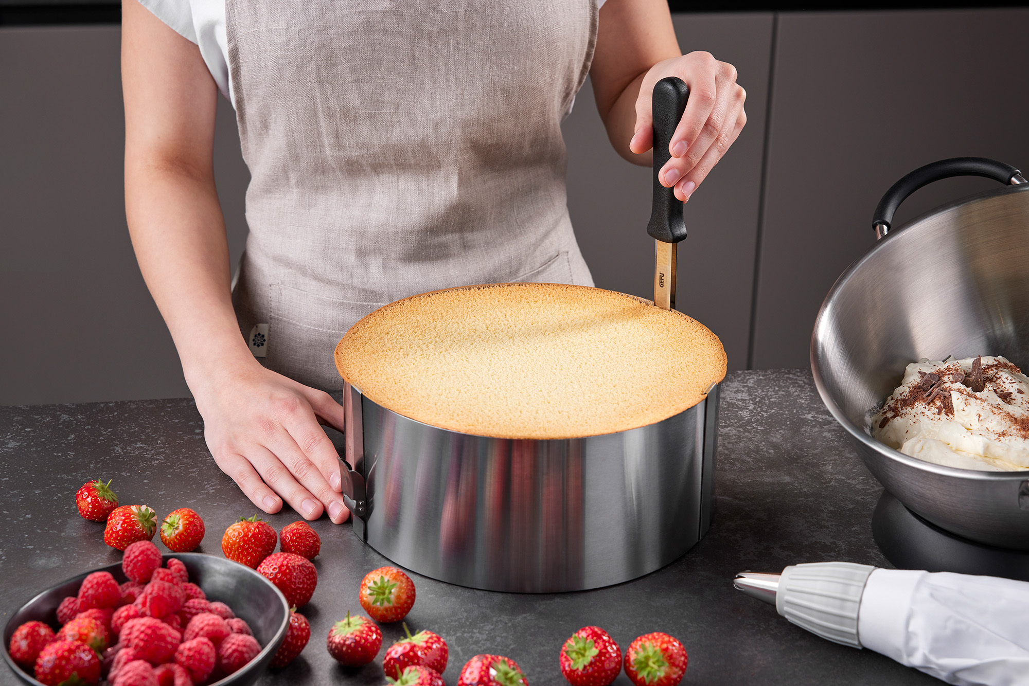 Kuchenbodenverarbeitung mit frischen Beeren und Sahne Eine Person schneidet einen Kuchenboden in einer Backform. Auf dem Tisch liegen Erdbeeren, Himbeeren, eine Schüssel mit Sahne und eine Spritztüte.