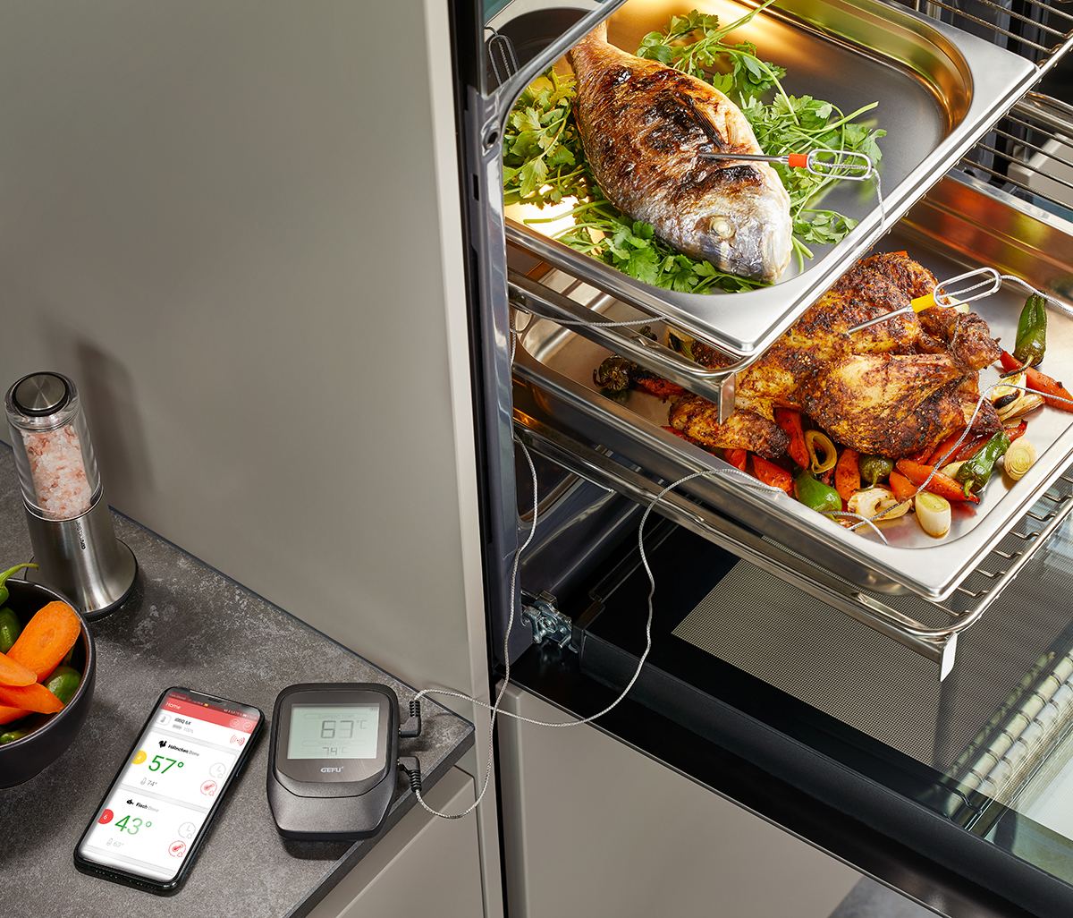 Offener Backofen mit einem Fisch auf Salat und einem gewürzten Hähnchen auf Gemüse. Daneben ein digitales Thermometer zur Überwachung der Kerntemperatur.
