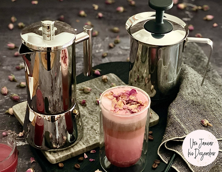 Pink Latte Zwei glänzende Espressokannen neben einem Glas mit cremigem Rosenkaffee, umgeben von Rosenblüten auf einem dunklen Hintergrund
