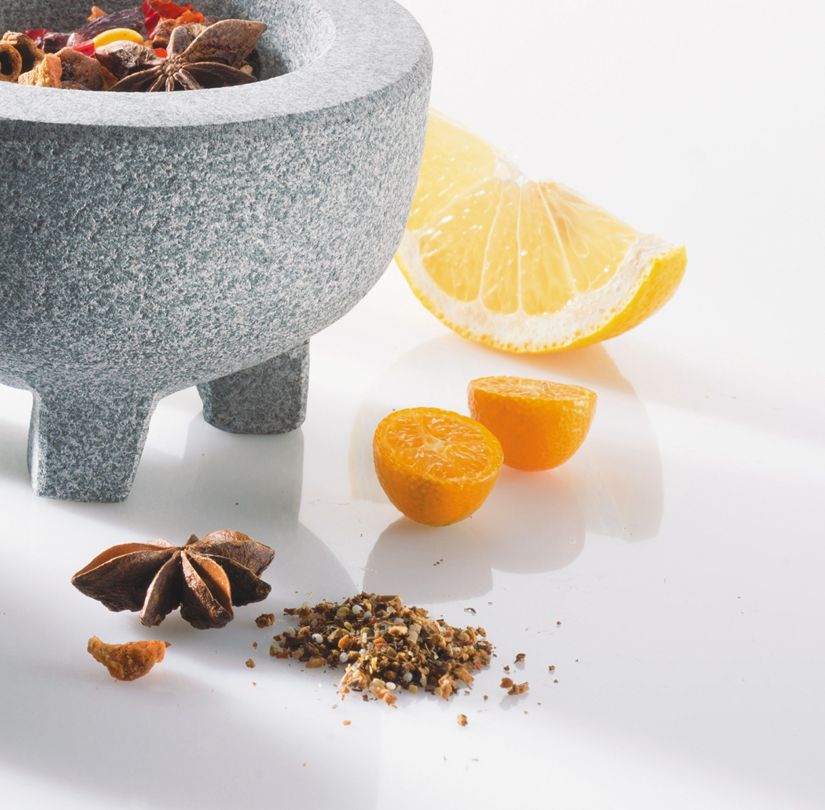 Steinmörser mit Gewürzen und Zitrusfrüchten Ein Steinmörser, umgeben von Sternanis, getrockneten Gewürzen und frischen Zitrusfrüchten auf einem weißen Hintergrund.