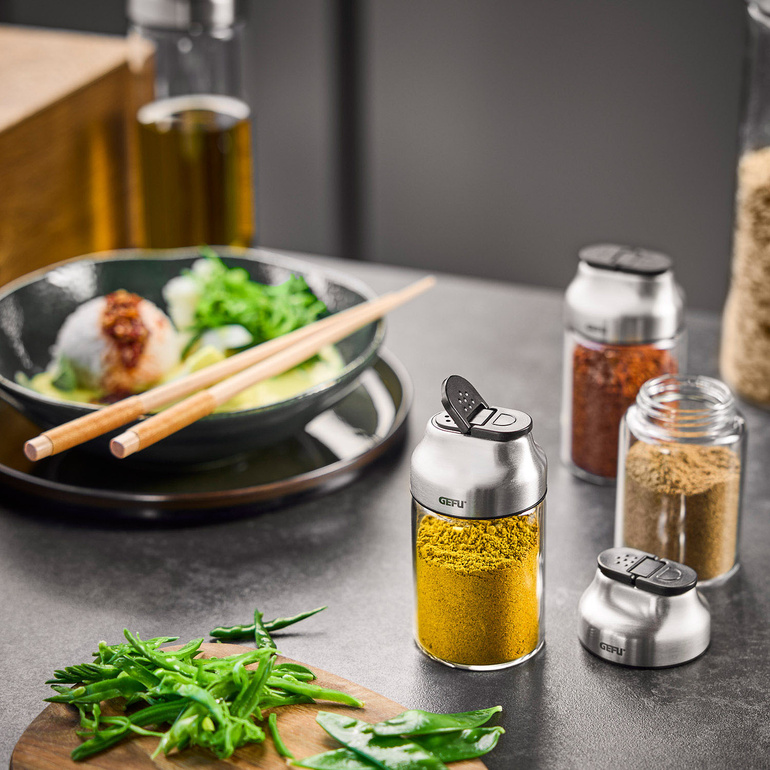Gewürzstreuer-Set aus Edelstahl in moderner Küche Ein Edelstahl-Gewürzstreuer-Set auf einer Küchenarbeitsplatte mit Curry, Pfeffer und Paprika, neben frischen Zutaten wie grünen Bohnen und einem Teller mit asiatischer Speise.