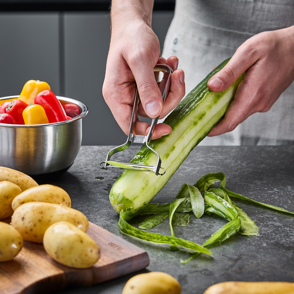 Detailaufnahme eines Gemüseschälers, der eine Gurke schält, mit einer Schüssel voller bunter Paprika und frischen Kartoffeln im Hintergrund auf einer Küchenarbeitsplatte