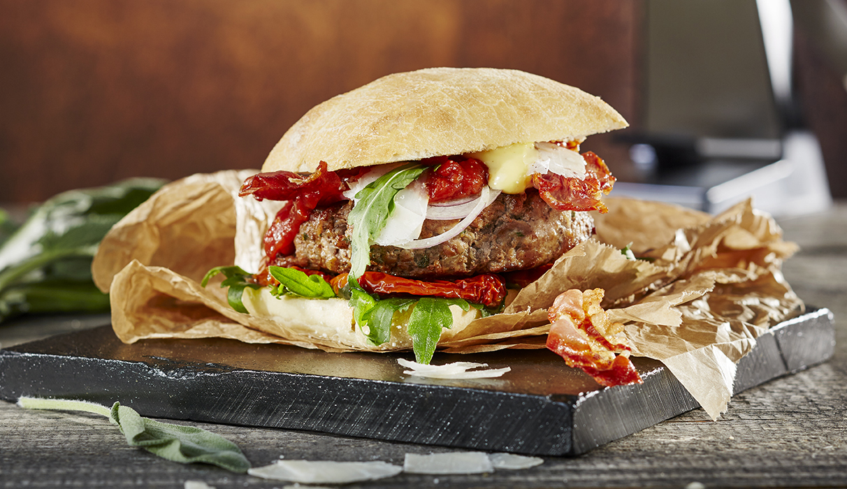Italien Style Kalbfleischburger Nahaufnahme eines Burgers mit Speck, Rucola, getrockneten Tomaten und gebratenen Zwiebeln auf einem rustikalen Holzbrett serviert