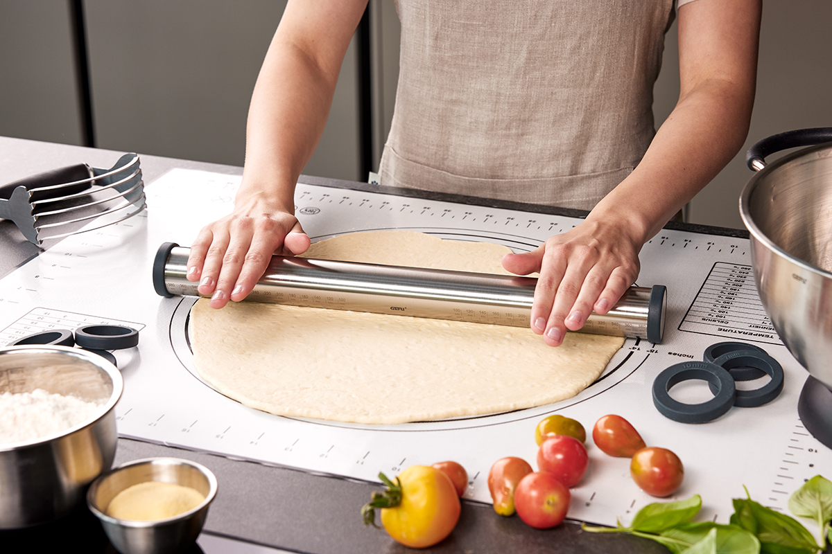 Professionelles Ausrollen von Teig auf einer Silikonmatte mit Edelstahl-Teigroller in einer modernen Küche Eine Person rollt Teig mit einem Edelstahl-Teigroller auf einer Silikonmatte aus; frische Tomaten und Mehl im Vordergrund. Perfekt für Backliebhaber.
